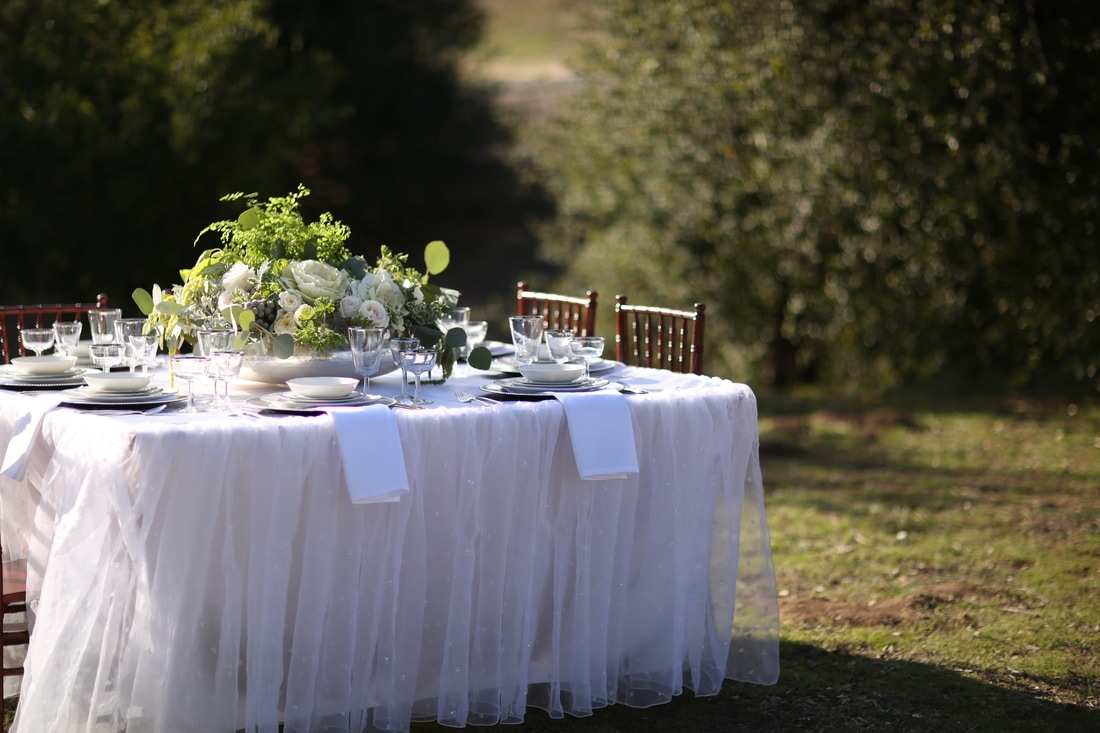 Square table with ruffled tablecloth. It is nestled in the oak trees at the outdoor venue in Murrieta.  The table is set with vintage china from Birdie in a Barn.