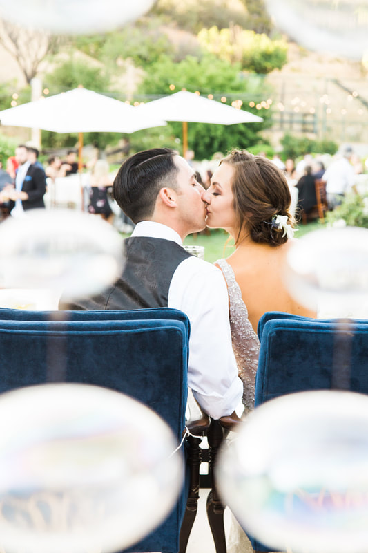 Bride and groom sneaking a kiss while seated in navy blue chairs at their sweetheart table.