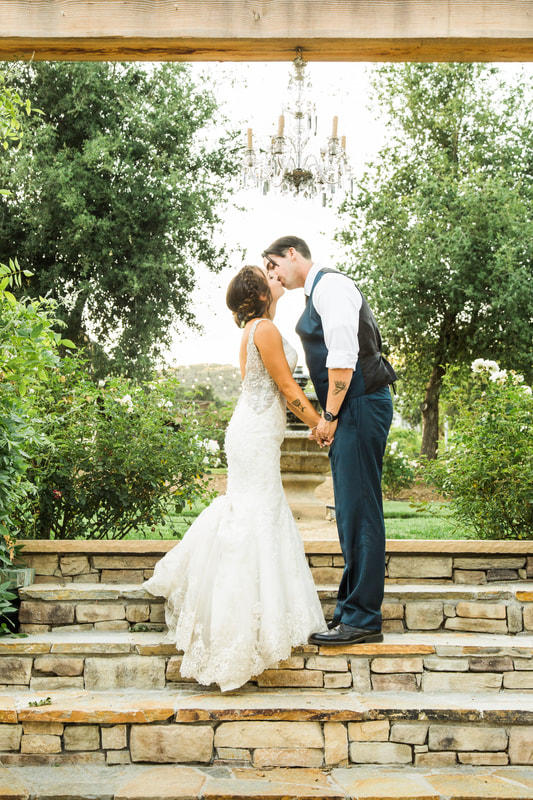 Bride and groom kissing under a chandelier hung from a wooden arch in this outdoor wedding venue.